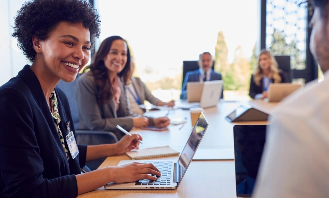 A group of people sit around a conference table with laptops and notepads, engaged in a meeting. One woman in the foreground is smiling.