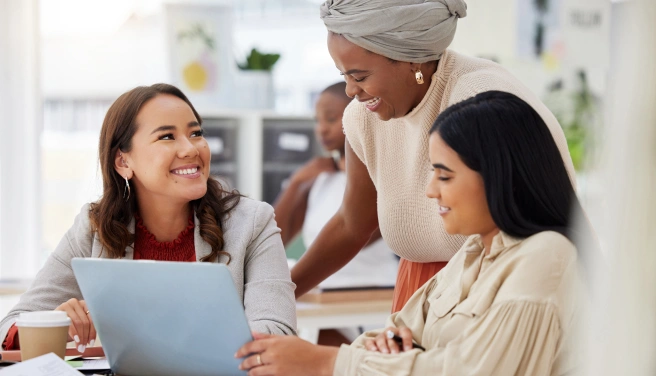 Three women sit and stand around a laptop, smiling and talking in a bright office setting.