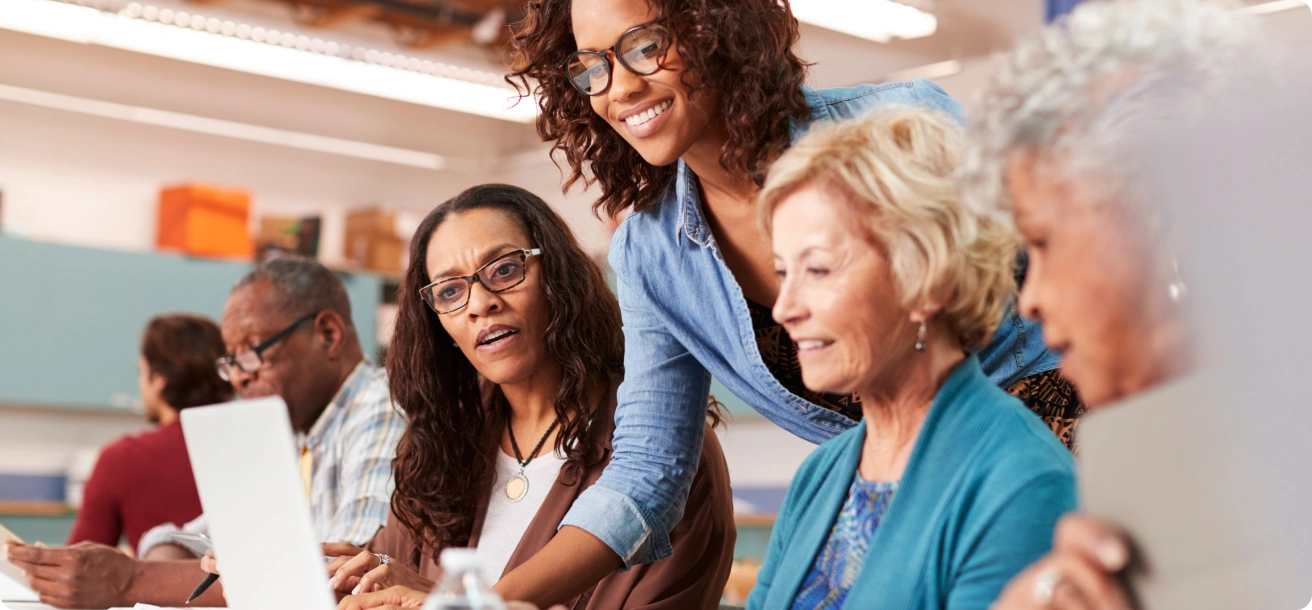 A group of adults sit together in a classroom, working on laptops, while a woman stands and assists them.