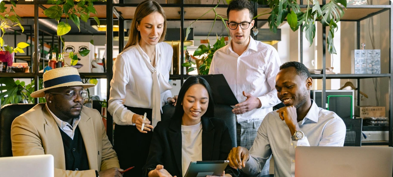 Five people in business attire are gathered around a table in an office, collaborating and discussing something on a tablet, with laptops and plants in the background.