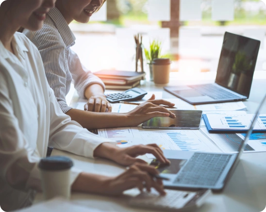 Three people work together at a desk with laptops, documents, and charts, focusing on data analysis. A coffee cup and office supplies are also visible.