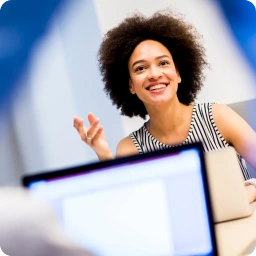 Woman with curly hair, wearing a striped sleeveless top, smiling and gesturing while seated at a desk with a laptop in an office setting.
