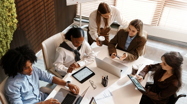 Five people sit around a table with laptops, tablets, and documents, collaborating on work in a modern office setting.