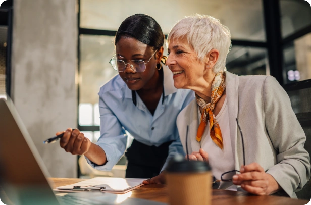 Two women, one younger and one older, look at a laptop screen together in an office setting, discussing documents with a coffee cup on the table.