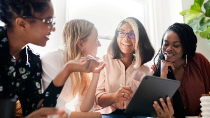 Four women are sitting together, smiling and talking while one holds a tablet. They appear to be having a discussion or collaborating in a bright indoor setting.