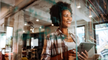 A woman stands indoors, smiling and holding a tablet, with graphs and charts visible on a glass wall in front of her.