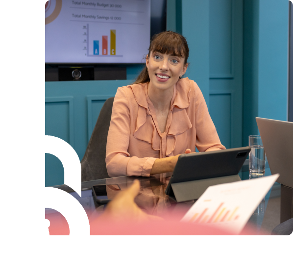 Woman in a pink blouse sits at a conference table with a tablet, engaging in a meeting; a presentation screen and laptops are visible in the background.